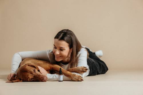 Frau liegt auf dem Boden und lächelt einen schlafenden Hund neben einer kleinen Flasche an. Neutraler Hintergrund.