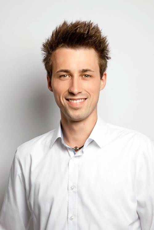 Lächelnder Mann mit stacheligen braunen Haaren, der ein weißes Hemd mit Knöpfen trägt, vor einem schlichten weißen Hintergrund.