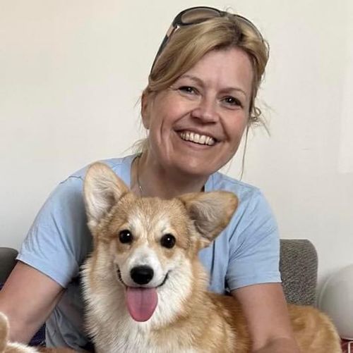 Lächelnde Frau mit Sonnenbrille auf dem Kopf, die auf einer Couch sitzt und einen fröhlichen Corgi mit heraushängender Zunge im Arm hält.