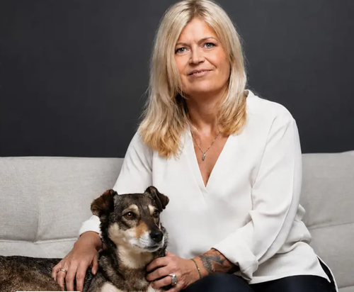 Eine Frau mit blonden Haaren in einer weißen Bluse sitzt auf einem Sofa, den Arm um einen braun-schwarzen Hund gelegt, vor einem dunklen Hintergrund.