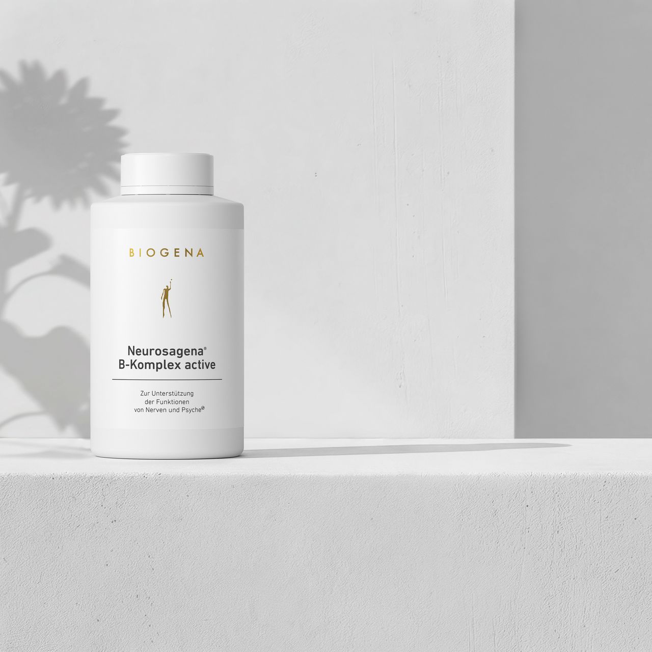 BIOGENA Neurosagena® B-Komplex active on a minimalist white shelf with soft shadows from a flower in the background.