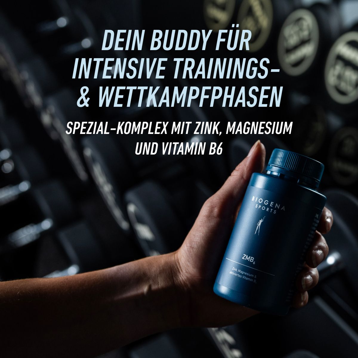 Ruka drží balenie BIOGENA SPORTS ZMB6 pred stojanom s činkami. Produkt je prezentovaný ako partner pre intenzívne tréningové a súťažné fázy s obsahom zinku, horčíka a vitamínu B6.