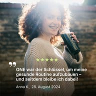Eine lächelnde Person hält einen BIOGENA ONE Drink in der Hand. Im Bild steht eine Kundenbewertung mit fünf Sternen: „ONE war der Schlüssel, um meine gesunde Routine aufzubauen – und seitdem bleibe ich dabei!“ von Anna K