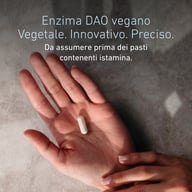 Una persona tiene una capsula nel palmo della mano. Il testo descrive BIOGENA DaoZym® Vegan, con un enzima DAO di origine vegetale, da assumere prima dei pasti che contengono istamina.