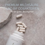 Mehrere weiße Kapseln liegen neben einer geöffneten BIOGENA Dose auf einer Steinoberfläche. Der Text beschreibt Premium-Milchsäure- und Bifidobakterien für ein gutes Bauchgefühl.